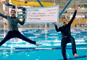 2 men jumping in a pool with a huge cheque