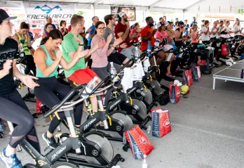 Large outdoor Cycle Class taking place under a tent with roughly 50 brightly dressed cyclists smiling and cheering