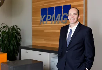 A professional shot of a man in a suite with light blue tie standing with his hands in his pockets smiling at the camera. There is a receptionist desk behind him with a sign that reads KPMG.