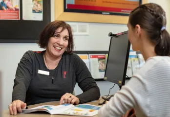 Middle aged woman with chin length dark hair in a grey YMCA shirt is sitting at a customer service desk in front of a computer and smiling at young female customer sitting in front of her.  