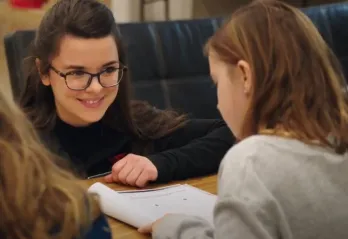 A young woman with glasses, brown hair and brown eyes is kneeling in front of a classroom table smiling at a 7 year old girl sitting at the table. The young girl has shoulder length brown hair, and her back is to the camera as she looks down at an open notebook in front of her.  