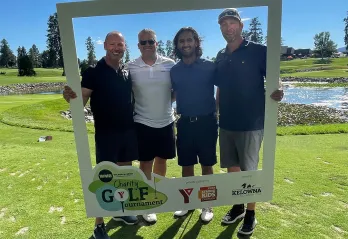 Four men in golf attire are standing outside on a golf course on a beautiful sunny day. They are all smiling at the camera and surrounded by a white frame they are holding around themselves. The men range in age from 30-55.
