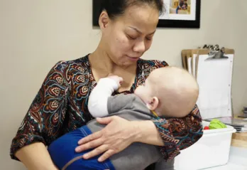 A woman in a patterned blouse is holding baby boy to her chest at a child care centre. The baby is asleep with his hand on her neck.