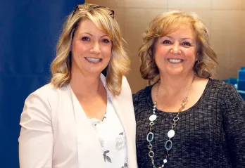 Two women are standing side by side and smiling at the camera. Allyson Graf is on the left with mid length blonde hair and a cream jacket, Sharon Peterson is on the right with shoulder length auburn hair and a grey sweater. 