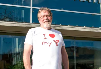 A man with glasses and a grey goatee is standing in front of the Kelowna Family Y and smiling at the camera. He’s wearing a white shirt that says ‘I love my Y’.