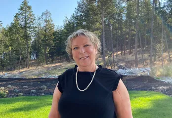 A woman with short blonde hair, wearing a black dress and pearls is smiling at the camera. There is a forest behind her and the sun is shining. 