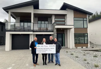 There are four adults standing in the driveway in front of a brand new modern luxury home. They are all smiling at the camera and holding a donation cheque of $150,000 to the KGH Foundation and YMCA of Southern Interior BC.