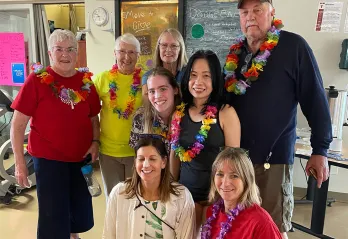 A group of 8 seniors and adults are standing together in a gym smiling at the camera. They are all wearing athletic clothes and ‘tourist wear’ including Hawaiian lays, tropical shirts and bright colors. 
