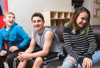 Three teens are sitting on a couch, smiling in different directions as they interact with people off camera. Two on the left are boys and there is one girl on the right.