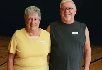 An older woman and man are standing in a gymnasium with their arms around one another smiling at the camera. The woman has grey hair and a yellow t-shirt while the man has white hair, glasses, and a grey sleeveless workout shirt on. 