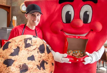 A YMCA mascot is holding an open box of smile cookies and posing beside a young female Tim Hortons Store Owner who is dressed as a large cookie. 