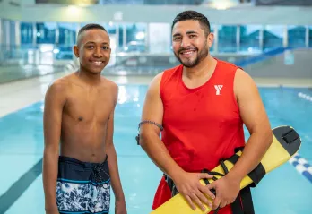 A young teen in swim trucks stands beside a male lifeguard with dark hair in front of a pool. They are smiling at the camera. 