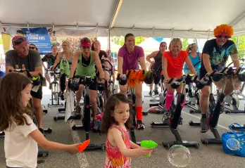 Two young girls with brown hair are playing with water guns and bubbles. Behind them a group of 14 adults dressed in costumes and athletic wear are riding cycle bikes in an outdoor tent and smiling. 