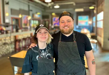 A young man and woman in black striped chef aprons are standing in a restaurant with their arms around one another and smiling at the camera. 