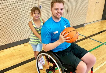 A young blonde man in a blue shirt and a wheelchair is smiling at the camera and holding a basketball in a gymnasium. There's an 8 year old girl holding the back handles of the chair also smiling at the camera. 