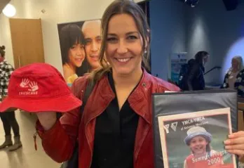 A young professional woman with dark hair is smiling at the camera and holding an older YMCA child’s bucket hat in one hand, and a photo of herself as a child in an old YMCA program advertisement in the other.