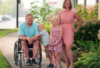 A family of 4 with two young children under the age of 7 are standing together outside in the sunshine and smiling at the camera.