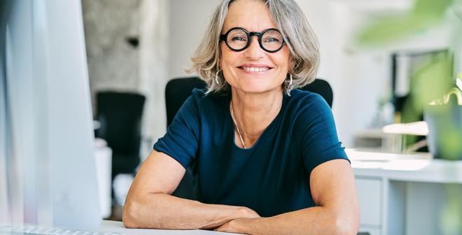 An older woman in a blue shirt with short grey hair and round black glasses is sitting at a desk in an office setting with her arms folded and smiling at the camera.