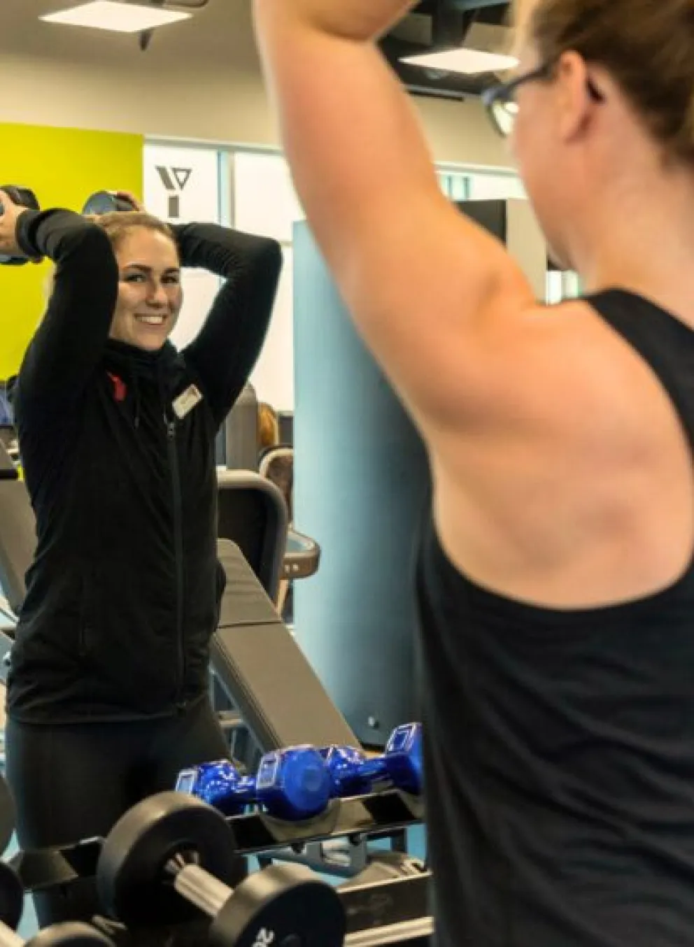 Two young women in black workout gear are standing in front of a mirror at the gym. They are both smiling and holding a large weight behind their head.