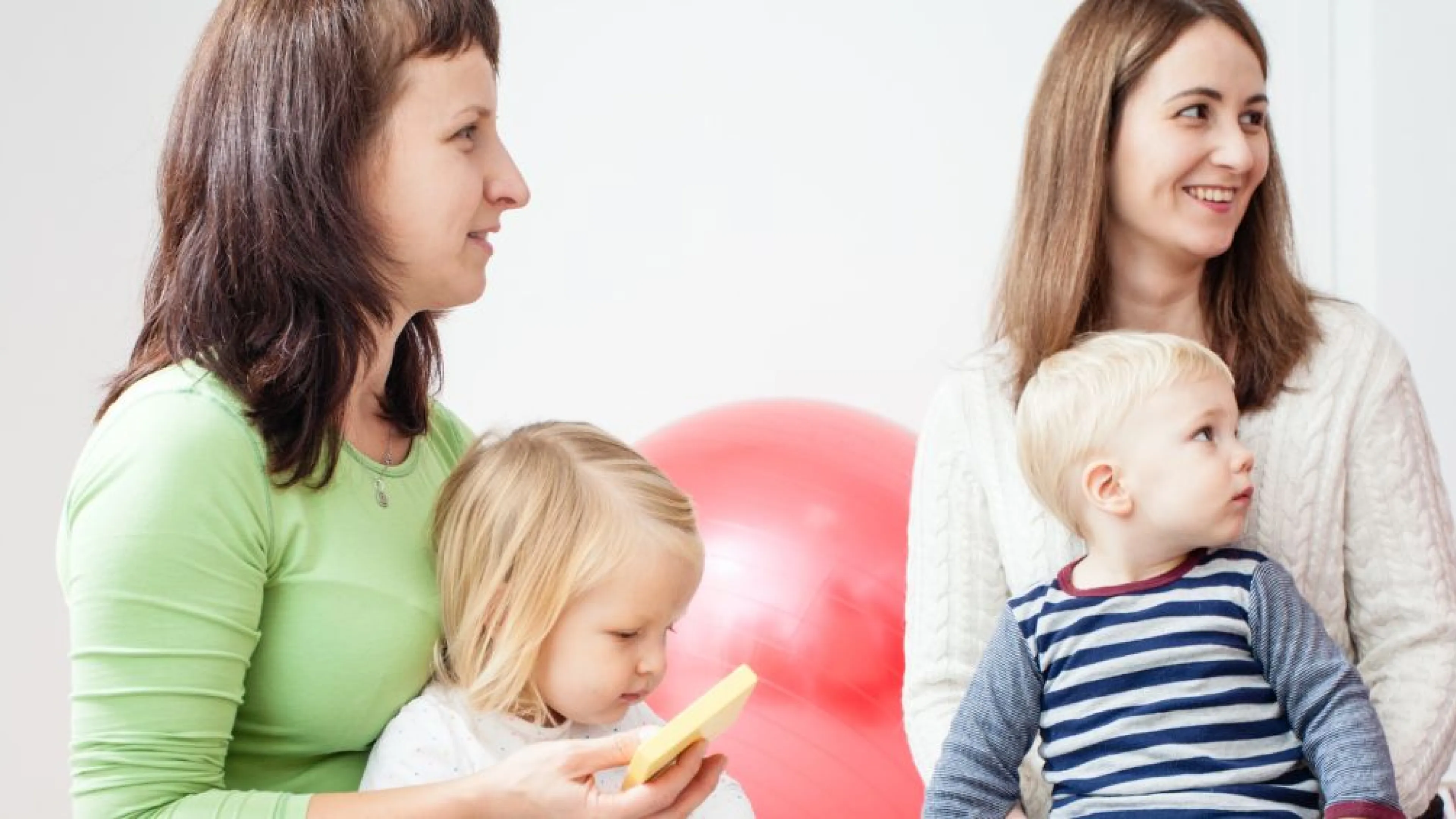 A group of young moms are sitting in a play room with their 3 year old children on their laps. They are smiling and listening to a speaker who is off screen.