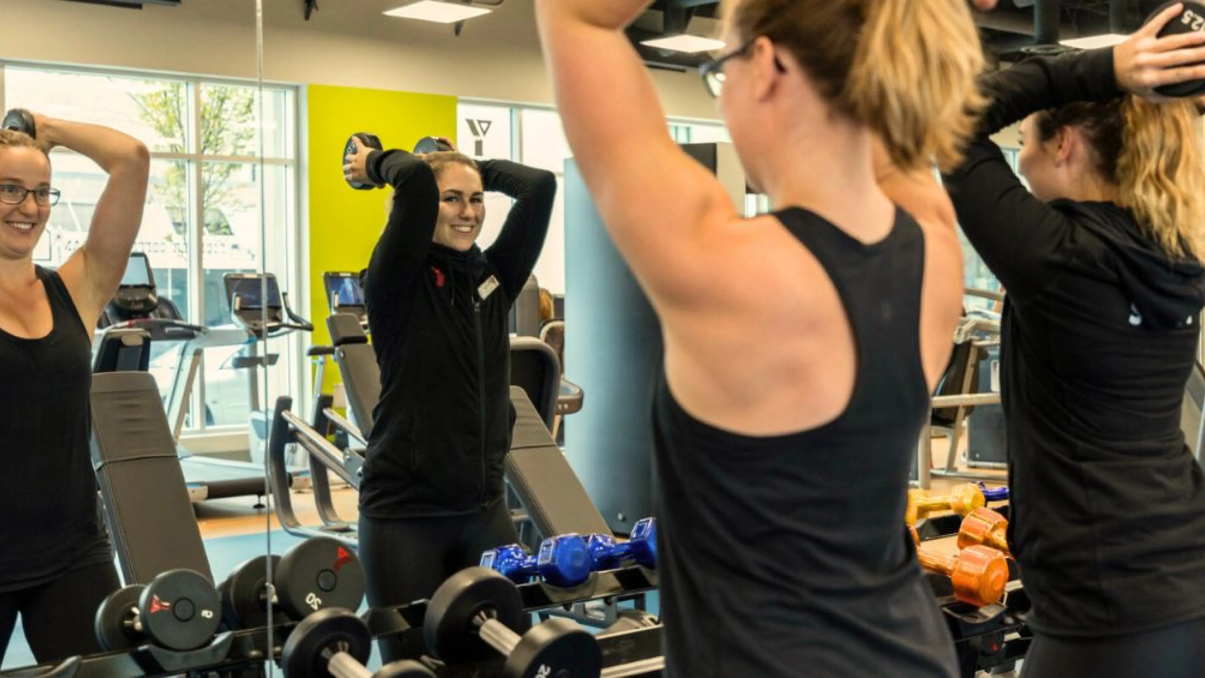 Two young women in black workout gear are standing in front of a mirror at the gym. They are both smiling and holding a large weight behind their head.