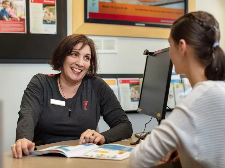 Middle aged woman with chin length dark hair in a grey YMCA shirt is sitting at a customer service desk in front of a computer and smiling at young female customer sitting in front of her.  
