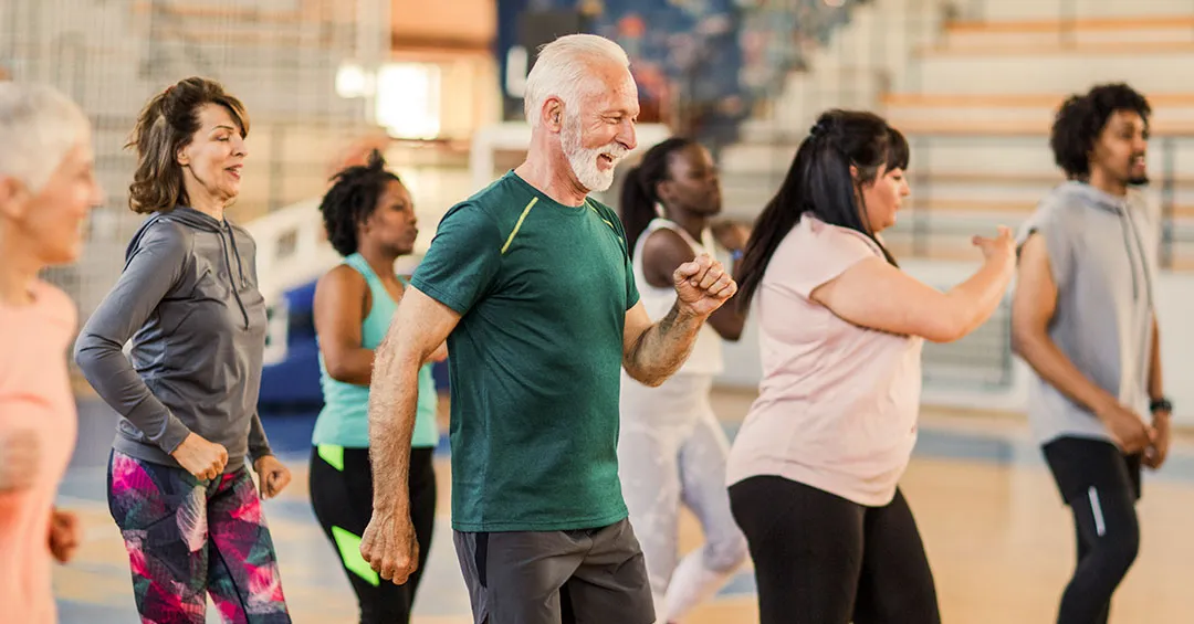 Seven adults in a group fitness class all mid step with arms swinging. There is an older gentleman in front with a green shirt and grey hair smiling.