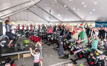  An outdoor Cycle class is under a large tent with dozens of participants dressed up brightly and cheering. There are bubbles blowing around the class and two children dancing in the foreground with one Cycle instructor on the a stage to the left leading the class. 