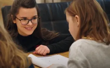 A young woman with glasses, brown hair and brown eyes is kneeling in front of a classroom table smiling at a 7 year old girl sitting at the table. The young girl has shoulder length brown hair, and her back is to the camera as she looks down at an open notebook in front of her.  