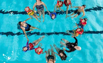 15 community members of all ages from senior to child, holding hands in a pool and looking up at the camera. Their bodies are forming the shape of a heart. 