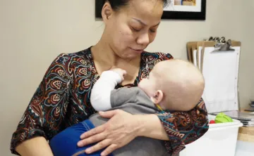 A woman in a patterned blouse is holding baby boy to her chest at a child care centre. The baby is asleep with his hand on her neck.