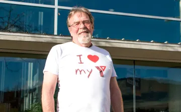 A man with glasses and a grey goatee is standing in front of the Kelowna Family Y and smiling at the camera. He’s wearing a white shirt that says ‘I love my Y’.