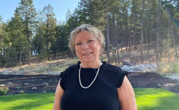 A woman with short blonde hair, wearing a black dress and pearls is smiling at the camera. There is a forest behind her and the sun is shining. 