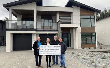 There are four adults standing in the driveway in front of a brand new modern luxury home. They are all smiling at the camera and holding a donation cheque of $150,000 to the KGH Foundation and YMCA of Southern Interior BC.