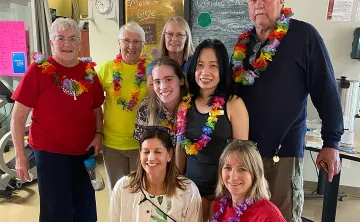 A group of 8 seniors and adults are standing together in a gym smiling at the camera. They are all wearing athletic clothes and ‘tourist wear’ including Hawaiian lays, tropical shirts and bright colors. 