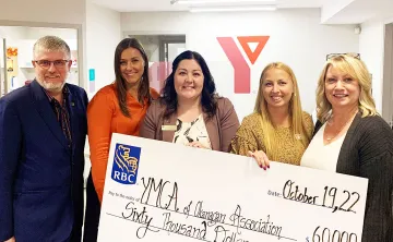 5 professionals of various ages in business clothing are standing in a YMCA office, holding a large donation cheque and smiling at the camera. 