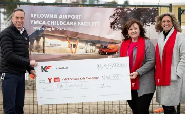 A man in a black jacket, and 2 women in grey winter coats and red YMCA scarves are all standing smiling at a camera holding a large cheque. In the background there is a construction site with a sign on the fence that reads ‘Kelowna Airport YMCA Childcare facility’.