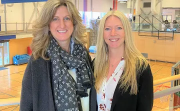 Two blonde women in professional clothing are standing in front of a gymnasium and smiling at the camera. 