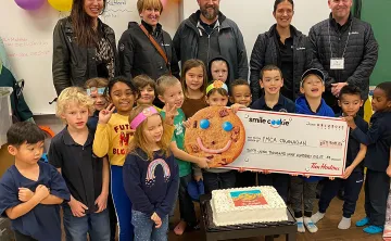 Five adults are standing in a classroom smiling at the camera. In front of them a group of 15 children aged 5-7 are smiling and holding a large donation cheque made out to the YMCA.