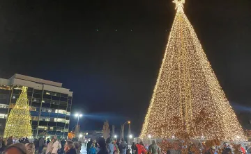 An enormous Christmas tree is lit up with all white lights  in the nighttime, as a crowd gathers around the base.  There are office buildings in the background that reflect the tree. 