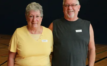 An older woman and man are standing in a gymnasium with their arms around one another smiling at the camera. The woman has grey hair and a yellow t-shirt while the man has white hair, glasses, and a grey sleeveless workout shirt on. 