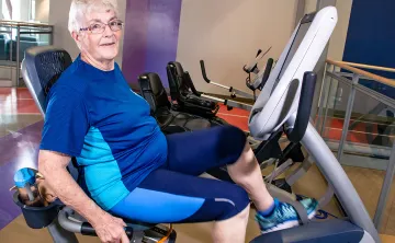 A woman with glasses, short white hair, and blue workout clothes is sitting on a laid back cycle machine in a gym and smiling at the camera. 