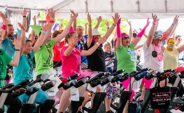 A group of 30 people are dressed up in costumes and bright colors, and sitting on stationary bikes in an outdoor class. They are all smiling and cheering and holding their hands up in the air. 