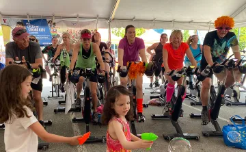 Two young girls with brown hair are playing with water guns and bubbles. Behind them a group of 14 adults dressed in costumes and athletic wear are riding cycle bikes in an outdoor tent and smiling. 