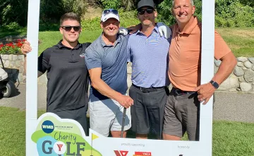 Four men in golf attire are holding clubs and smiling at the camera on a golf course.