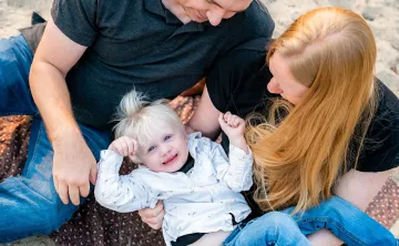A mother and folder are holding their 2 year old boy and looking down at him as they sit on a beach. The little boy has blue eyes, blonde hair, and is smiling up at the camera.