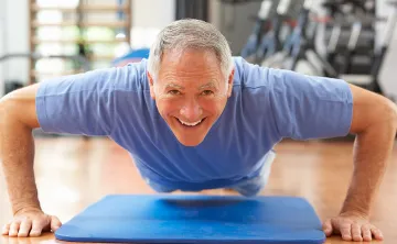 An older gentleman with grey hair and a blue t shirt is hovering in a pushup over a blue mat in a gym, and smiling at the camera. 