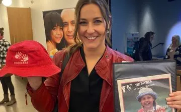 A young professional woman with dark hair is smiling at the camera and holding an older YMCA child’s bucket hat in one hand, and a photo of herself as a child in an old YMCA program advertisement in the other.
