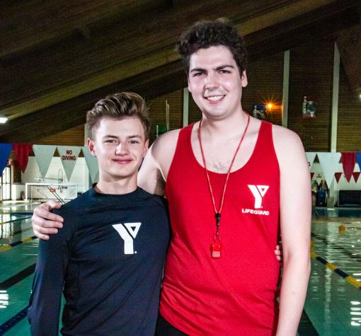Two male lifeguards standing on a pool deck. One in a red lifeguard tank has his arm around the other in a black YMCA rashguard. They are both smiling at the camera.