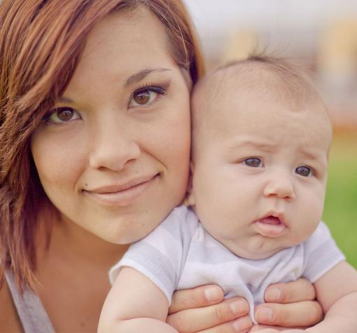 A close up photo of a young mother with brown eyes and dark red hair holding a three month old baby in a white shirt.
